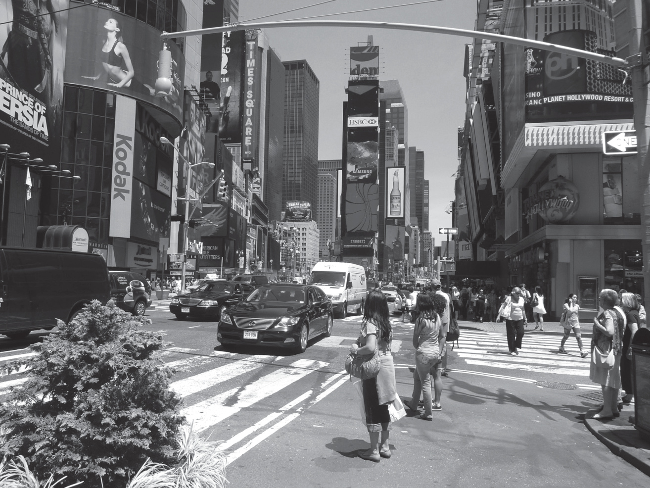 Photographie en noir et blanc d'un carrefour de Times Square à New York : taxis, panneaux publicitaires (Kodak, Adopt) et piétons à un passage clouté.