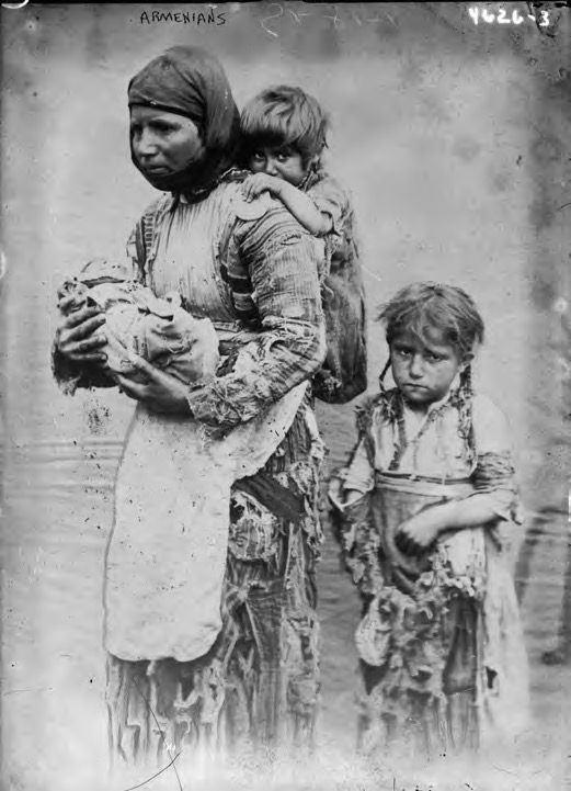 A black-and-white period photograph showing an Armenian mother carrying a young child on her back, followed by another child, walking along a path.