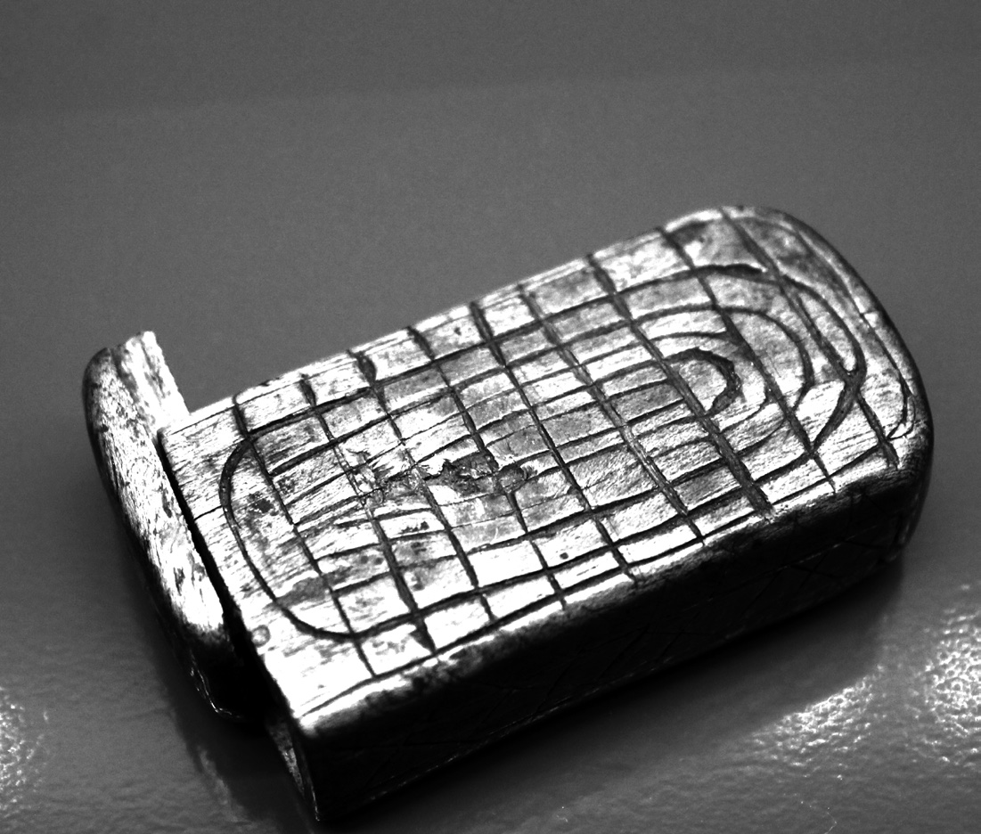 Fotografía en blanco y negro de una mezuzá portátil de metal labrado, colocada sobre una tela oscura, vista de tres cuartos.